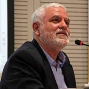 Foto de Rudá Ricci. Na imagem, ele está com cabelo e barba grisalhos. Ele tem uma expressão séria e contemplativa, olhando ligeiramente para cima. Ele está vestido com um blazer escuro sobre uma camisa azul clara. Ele está sentado em um ambiente de conferência, com um microfone à sua frente e uma projeção visível ao fundo.