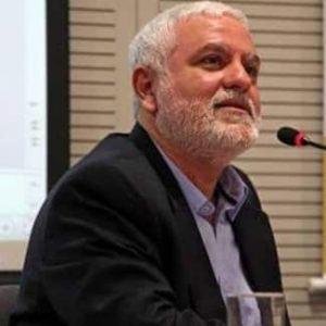 Foto de Rudá Ricci. Na imagem, ele está com cabelo e barba grisalhos. Ele tem uma expressão séria e contemplativa, olhando ligeiramente para cima. Ele está vestido com um blazer escuro sobre uma camisa azul clara. Ele está sentado em um ambiente de conferência, com um microfone à sua frente e uma projeção visível ao fundo.